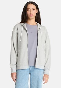 Hoodie zippé gris clair avec cordons de serrage, présentant une coupe décontractée et des bords côtelés, superposé sur un T-shirt violet clair. Jeans en denim visibles.