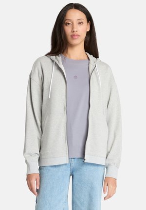 Hoodie zippé gris clair avec cordons de serrage, présentant une coupe décontractée et des bords côtelés, superposé sur un T-shirt violet clair. Jeans en denim visibles.