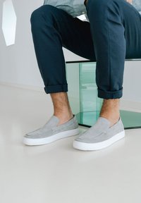 Graue Wildlederschuhe zum Hineinschlüpfen mit einer weißen Gummisohle, die eine glatte Textur und ein minimalistisches Design aufweisen. Getragen mit hochgekrempelten dunkelblauen Hosen.