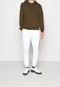 Brauner Kapuzenpullover mit schwarzen Kordeln und rotem Logo auf der Brust, kombiniert mit weißen Trainingshosen mit rotem Text an der Seite.