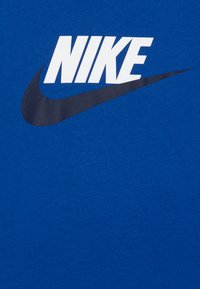 Tricou din bumbac albastru, cu un mare logo alb Nike și un design de swoosh de un albastru închis. Tesătura pare netedă, cu o ușoară textură.