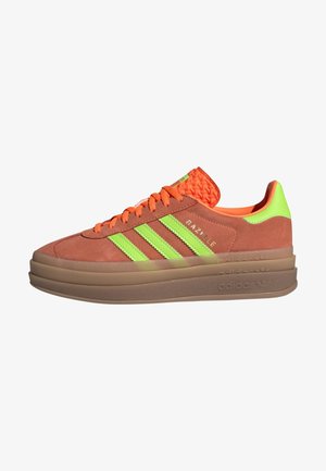 Bruine suède Adidas Gazelle sneaker met neon groene strepen, oranje veters en tong, en een dikke bruine rubberen zool.