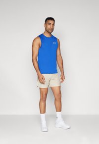 Modrá bezrukávová sportovní trika, béžové šortky s prodyšnou strukturou, bílé tenisky a ponožky do kotníku. Logo Under Armour na tričku a šortkách.