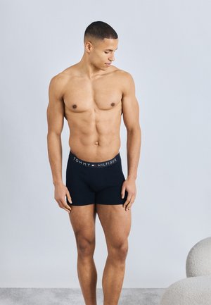 Μυώδης άντρας, ξυπόλητος, φοράει μαύρα μποξεράκια Tommy Hilfiger, κοιτάζοντας προς τα κάτω και αριστερά, με φόντο ένα απλό φωτεινό υπόβαθρο.