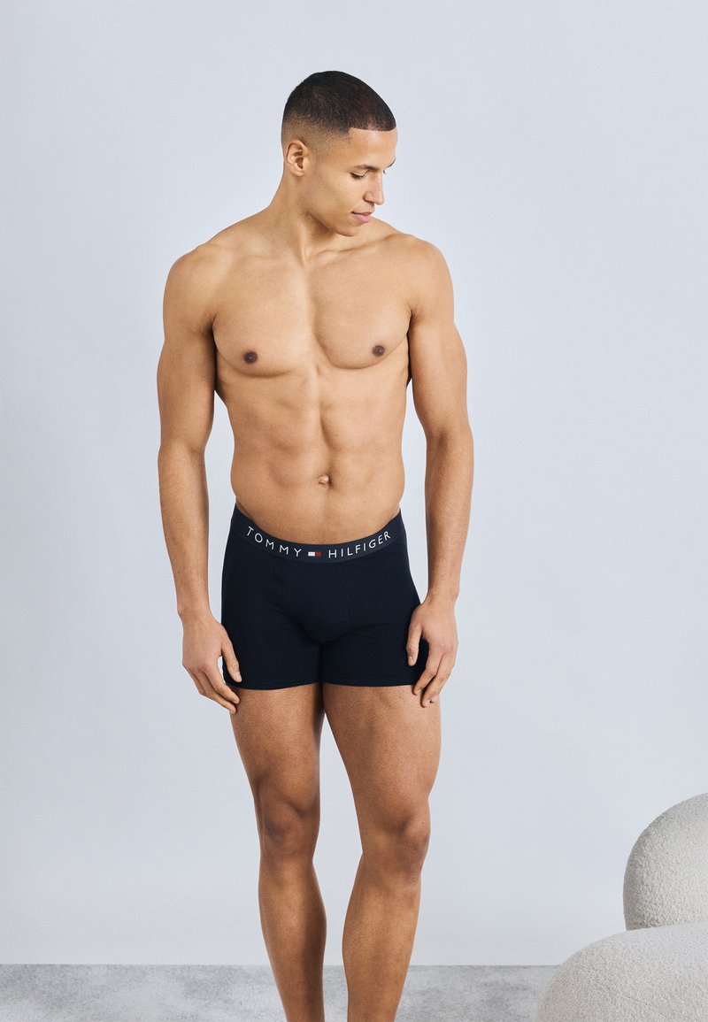 Μυώδης άντρας, ξυπόλητος, φοράει μαύρα μποξεράκια Tommy Hilfiger, κοιτάζοντας προς τα κάτω και αριστερά, με φόντο ένα απλό φωτεινό υπόβαθρο.