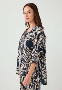 Blusa fluida a maniche lunghe di colore blu navy con un motivo astratto beige. Tessuto leggero, colletto e spacco laterale per facilitare i movimenti.