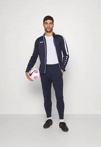 Marineblauer sportlicher Trainingsanzug mit weißen Streifen, bestehend aus einer Jacke mit Reißverschluss und enganliegenden Hosen. Das Modell hält einen gemusterten Fußball und trägt Fußballschuhe.