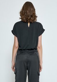 Blusa nera a maniche corte con dettagli a oblò sul retro, realizzata in tessuto liscio, abbinata a pantaloni cargo scuri con accentuazioni di tasche.