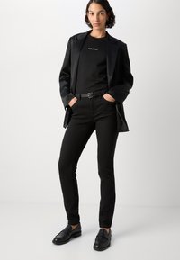 Zwarte op maat gemaakte blazer over een zwart T-shirt met logo, gecombineerd met zwarte slim-fit broeken en zwarte loafers. Gladde textuur, minimalistisch ontwerp.