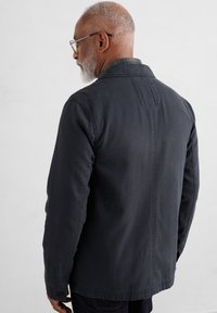 Jachetă bleu închis cu guler, având un material texturat și o croială dreaptă. Prezintă un model subtil de dungi orizontale pe spate.