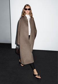 Brauner Wollmantel ohne Kragen, kombiniert mit einem weißen Rollkragenpullover und schwarzen Leggings. Eine braune Tasche und dunkle Sonnenbrillen runden den Look ab.