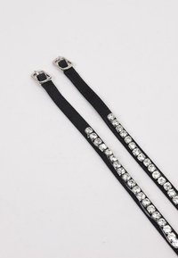 Tirantes de cuero negro con herrajes plateados y adornos de pedrería a lo largo de un lado. Textura suave, dos cierres de hebilla ajustables.