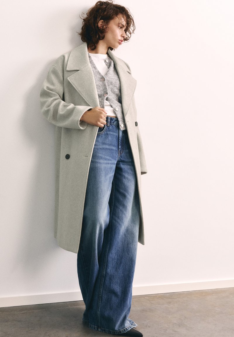 Femme portant un long manteau gris clair, un cardigan gris, un haut blanc et un jean large bleu, debout contre un mur blanc.