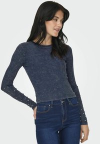 T-shirt a maniche lunghe a coste in blu scuro con leggeri puntini; presenta dettagli in perle sulle maniche; abbinato a jeans blu.