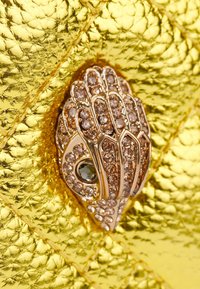 Superficie dorata texture con un motivo decorativo a forma di mano dorata, impreziosita da pietre scintillanti e da un gemma verde al centro.