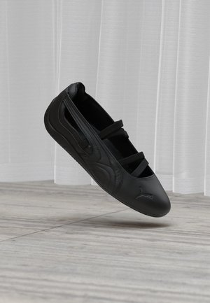 Black athletic slip-on shoe with three elastic straps, Puma logo on toe, shown mid-air above light wood floor and white curtains.