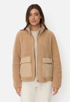 Kvinde iført en tan sherpa fleecjakke med frontlynlås og to store klaplommer, kombineret med en let sweater og hvide bukser.