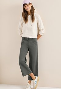 Mujer sonriente con sudadera ligera, pantalones anchos gris cortos, zapatillas blancas con detalles en azul y gorra morada sobre fondo beige.