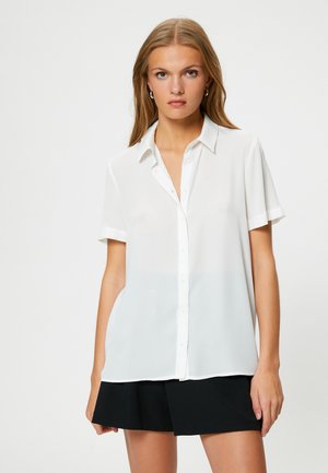 Camicia bianca a maniche corte con bottoni, realizzata in tessuto leggero. Presenta un colletto classico e una vestibilità rilassata, abbinata a una gonna nera.