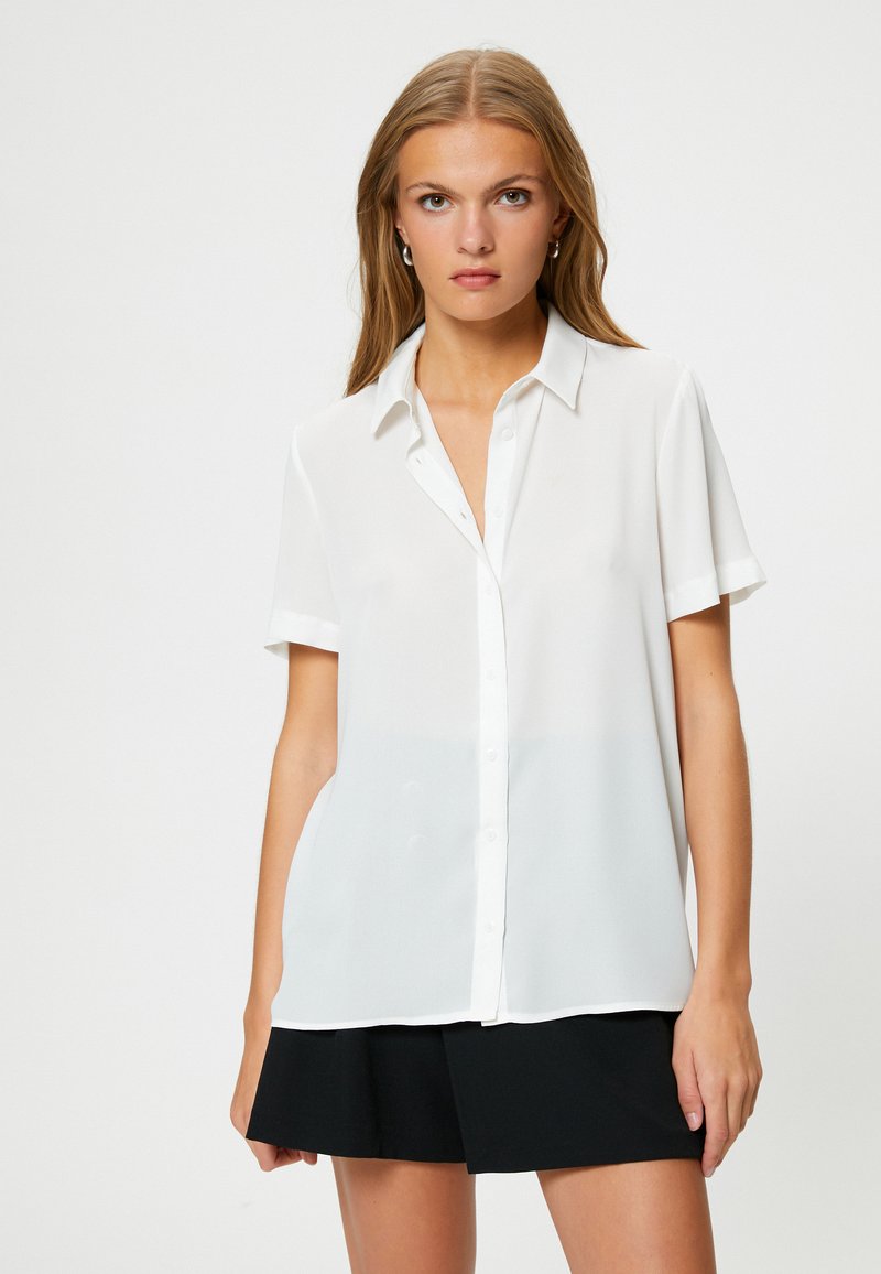 Camicia bianca a maniche corte con bottoni, realizzata in tessuto leggero. Presenta un colletto classico e una vestibilità rilassata, abbinata a una gonna nera.