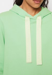 Jasnozielona bluza z kapturem wykonana z miękkiego materiału, z haftowanym logo na sznurku oraz luźnym krojem, wyposażona w kieszeń kangurkę.