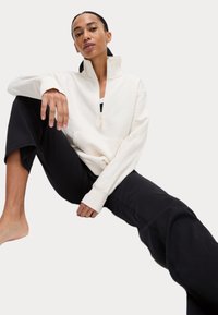 Krämfärgad zip-up sweatshirt med hög krage, ribbade mudd och en kangaroo-lomme, parat med svarta löst sittande byxor. Slät textur.