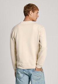 Felpa beige con polsini e orlo a costine, dotata di colletto rotondo e texture liscia. Indossata con jeans in denim azzurro chiaro.