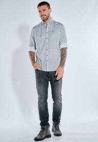 Man with tattoos wearing patterned button-up shirt with rolled sleeves, dark jeans, and gray lace-up boots, standing with one hand in pocket.