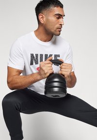 Svart kettlebell holdt av en person i en hvit Nike T-skjorte med en teksturert logo, kombinert med svarte treningbukser. Kort hår, sideprofil.