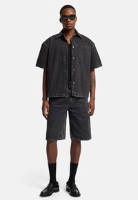Camicia nera di denim a maniche corte con tasca, abbinata a pantaloni corti in denim neri. Indossata con calze nere e mocassini, e occhiali da sole scuri.