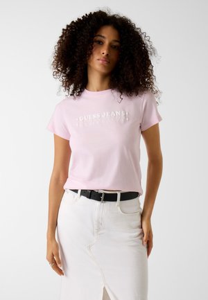 Frau mit lockigem Haar, die ein hellrosa "Guess Jeans" T-Shirt trägt, das in einen weißen Rock mit schwarzem Gürtel gesteckt ist, vor einem schlichten Hintergrund stehend.