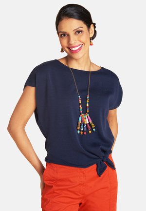 Lächelnde Frau mit zurückgebundenen dunklen Haaren, trägt ein marineblaues Oberteil mit seitlichem Knoten, orangene Hose sowie eine bunte Perlenkette und Ohrringe.