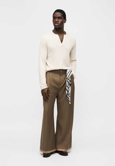 Άνδρας που φοράει κρεμ πουκάμισο με μακριά μανίκια, καφέ παντελόνι ψηλόμεσο με φαρδιά πόδια, σκούρα παπούτσια, κρατώντας ένα κασκόλ με σχέδια στην τσέπη.