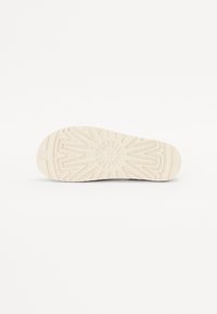 Sneakerin pohja, jossa on teksturoitu, vaaleanharmaa kumipinta, jossa on geometrinen kuvio ja pyöreä emblemipainatus kantapäässä.