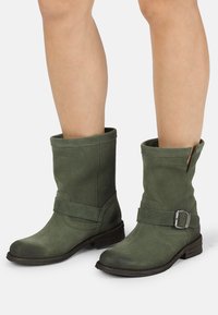 Botines de ante verde con un puño enrollado, tacón plano, cremallera lateral y una correa decorativa con detalle de hebilla plateada.