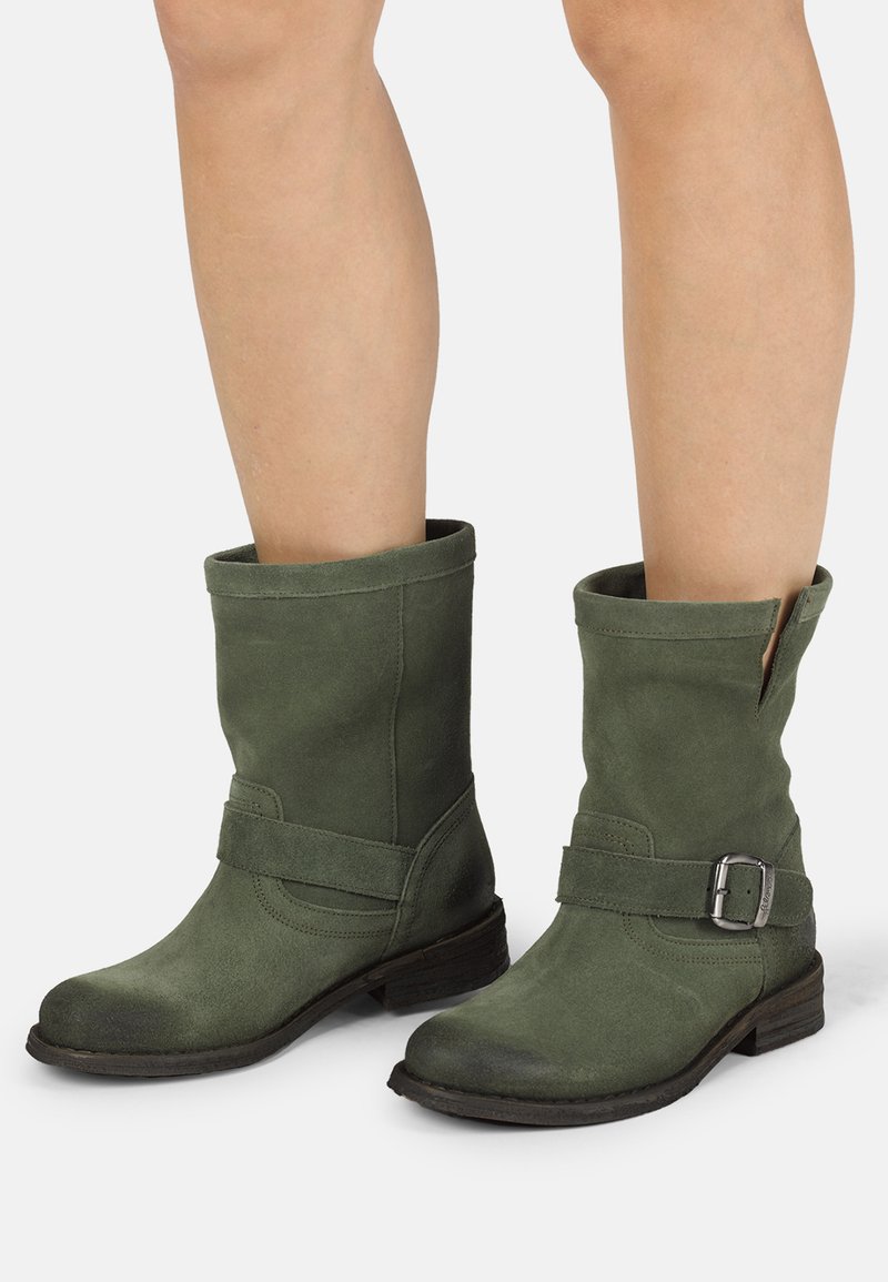 Botines de ante verde con un puño enrollado, tacón plano, cremallera lateral y una correa decorativa con detalle de hebilla plateada.