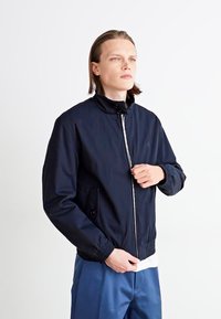 Chaqueta bomber azul marino hecha de tela suave; cuenta con cierre de cremallera al frente, cuello alto, puños elásticos y bolsillos laterales.