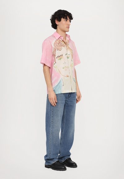 Camisa de manga corta, rosa, con un diseño de cara, con frente abotonado, corte relajado, combinada con vaqueros anchos azules y zapatos negros.
