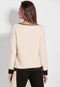 Maglia a maniche lunghe color crema con dettaglio nero sul colletto, vestibilità comoda e polsini a coste. Il materiale appare morbido e liscio con un design semplice.