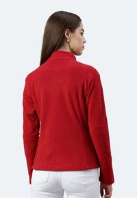 Giacca in pile rossa con colletto, maniche lunghe e design aderente. La texture sembra morbida, con uno stile minimalista.