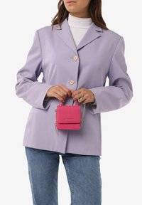 Lavendel blazer met lichtroze knopen, gecombineerd met een kleine, gestructureerde roze handtas met een kettingriem, vastgehouden voor lichtblauwe jeans.