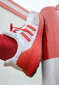 Bílé sportovní boty s červenými akcenty, texturovanou podešví a prodyšným síťovaným svrškem s výrazným designem tří pruhů. Viditelný červený nátepník.