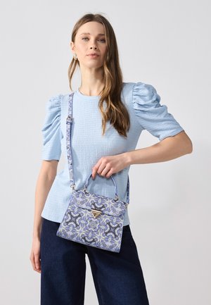 Vrouw draagt een lichtblauw topje met pofmouwen en een donkere broek, houdt een blauw patroonhandtasje met een driehoekige gesp vast.
