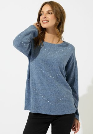 Lächelnde Frau mit schulterlangem braunem Haar, die einen lockeren blauen Pullover mit kleinen weißen Punkten und schwarze Hosen trägt, posiert mit der Hand nahe ihrem Haar.