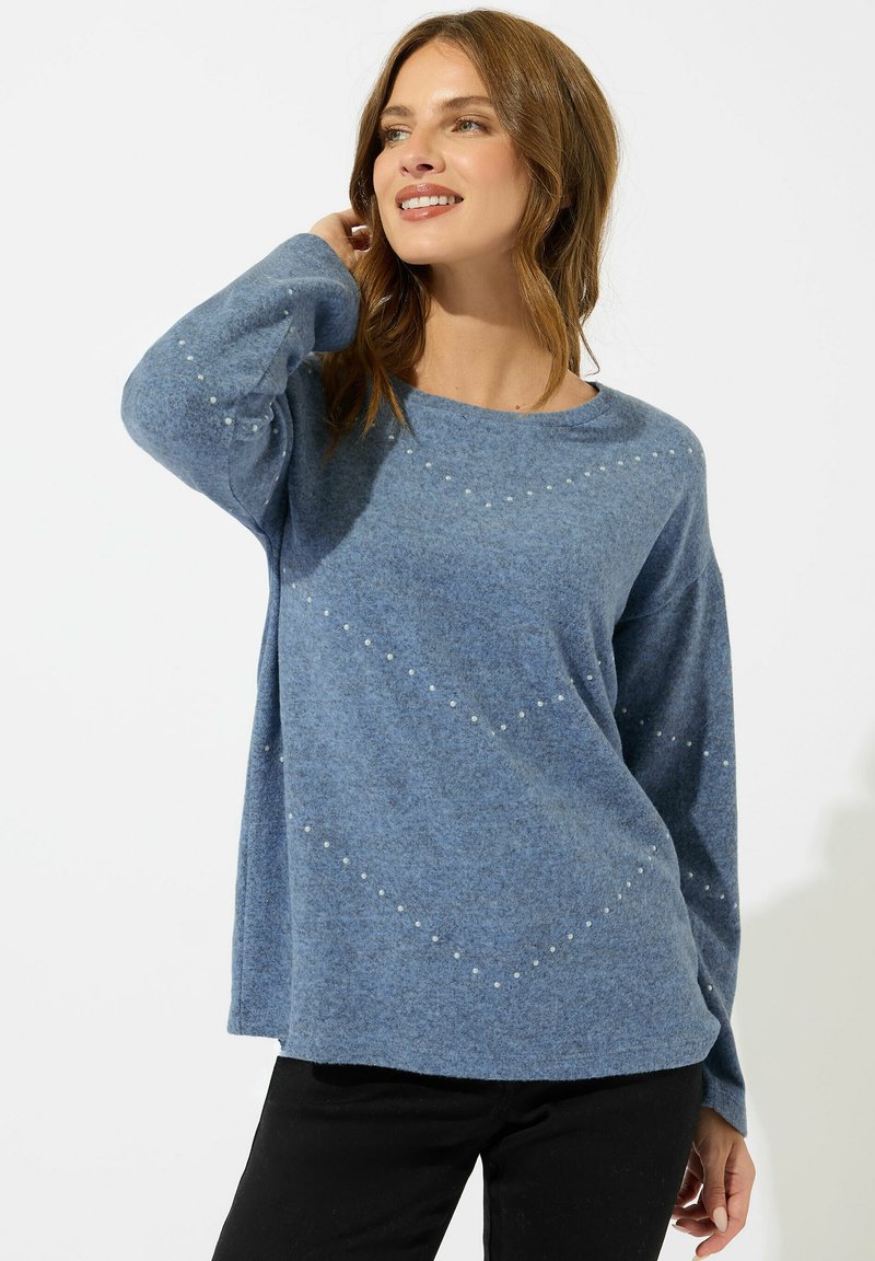 Donna sorridente con capelli castani lunghi fino alle spalle, che indossa un maglione blu svolazzante con piccoli motivi bianchi e pantaloni neri, in posa con la mano vicino ai capelli.