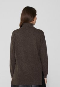 Suéter marrón con cuello alto, puños acanalados y una parte trasera ligeramente más larga. Textura suave con un ajuste relajado y aberturas a los costados.