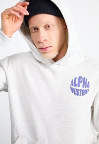 Šviesiai pilkas džemperis su purpuriniu apvaliu „ALPHA INDUSTRIES“ logotipu, su kengūros kišene ir reguliuojama kapuce. Tekstūruotas audinys.