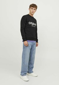 Sudadera negra con el gráfico de texto "Original" en el frente, combinada con jeans azul claros y zapatillas blancas, de pie contra un fondo liso.