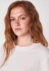 Een crèmekleurig, zacht t-shirt met een ronde halslijn, met een ontspannen pasvorm en een gladde textuur, gedragen door een persoon met golvend, rood haar.