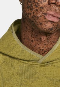 Hanorac verde măsliniu cu țesătură texturată și un design patternat. Include o glugă cu șnur și un tiv ușor contrastant în jurul gâtului.